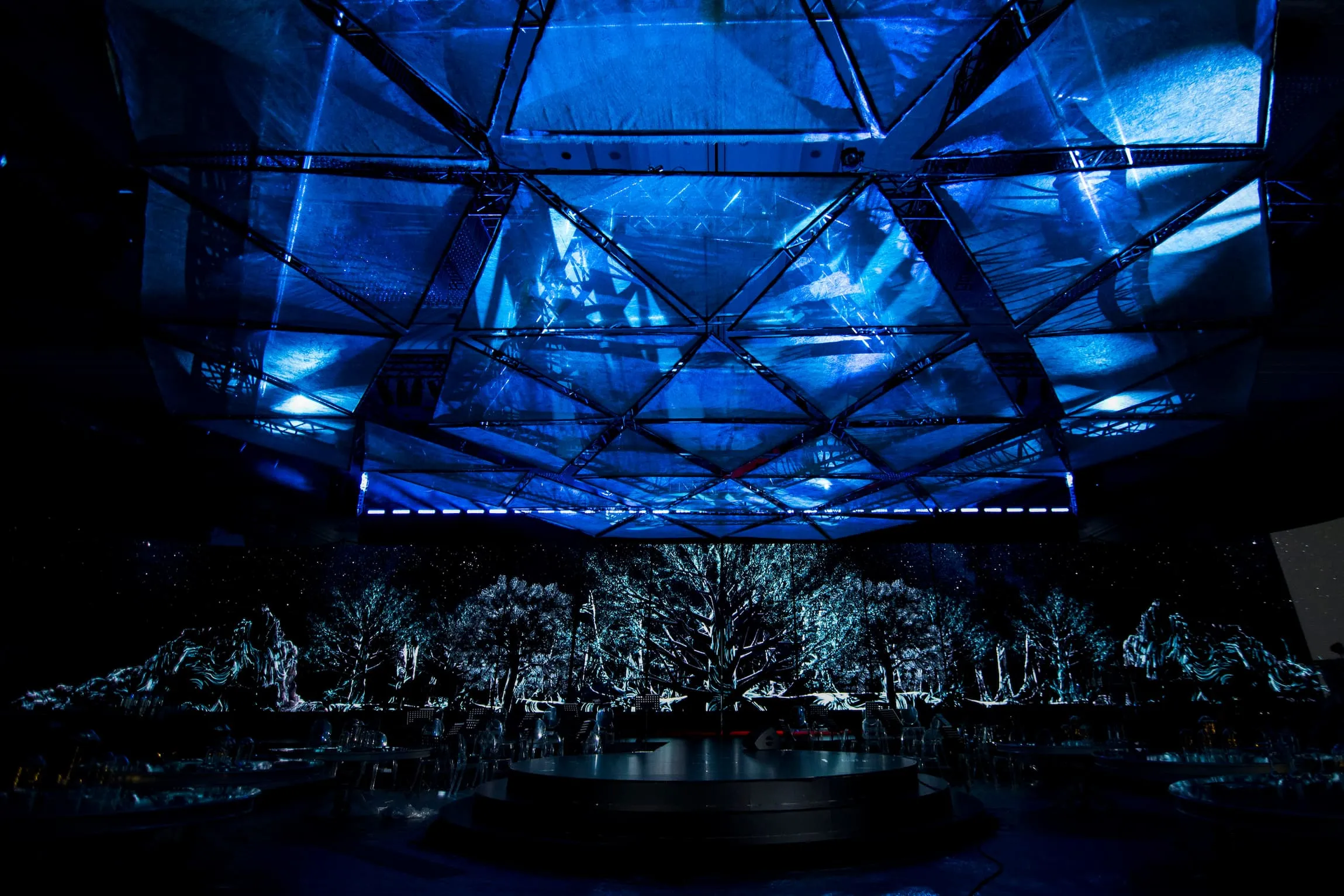 The 360° stage lit blue under a geometric canopy, silver tree-like forms glowing around its base