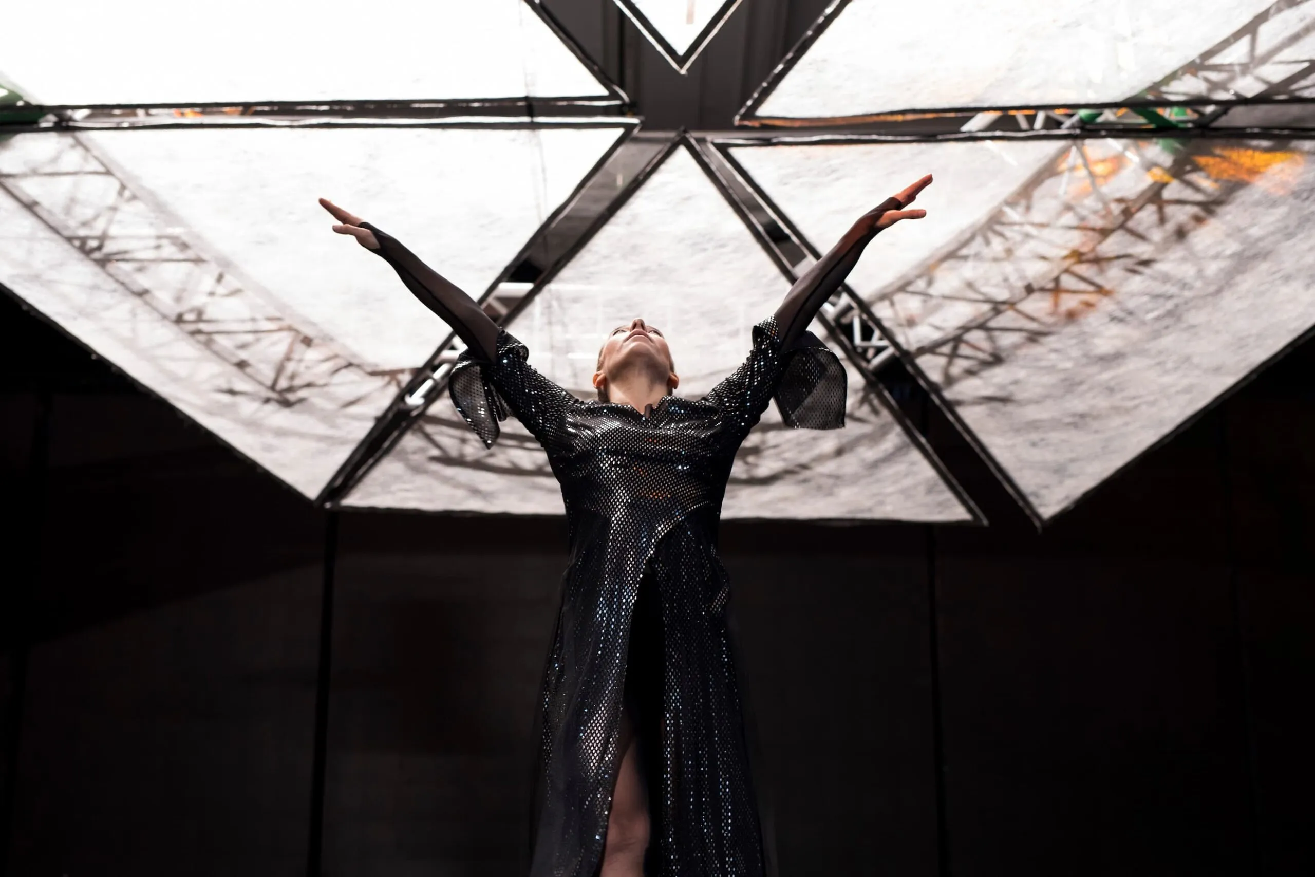 A dancer in a black sequinned gown raises both arms beneath a suspended triangular canopy of light