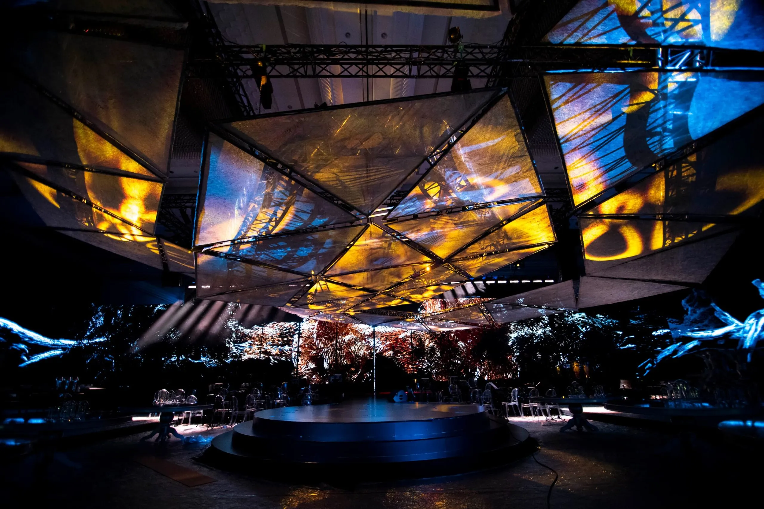 The 360° stage under a canopy of triangular screens glowing gold and blue, tables set around the round platform