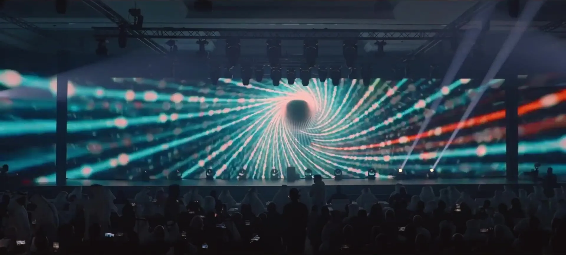 A vortex of light lines spins across the wide stage screen as lasers cut over the seated audience