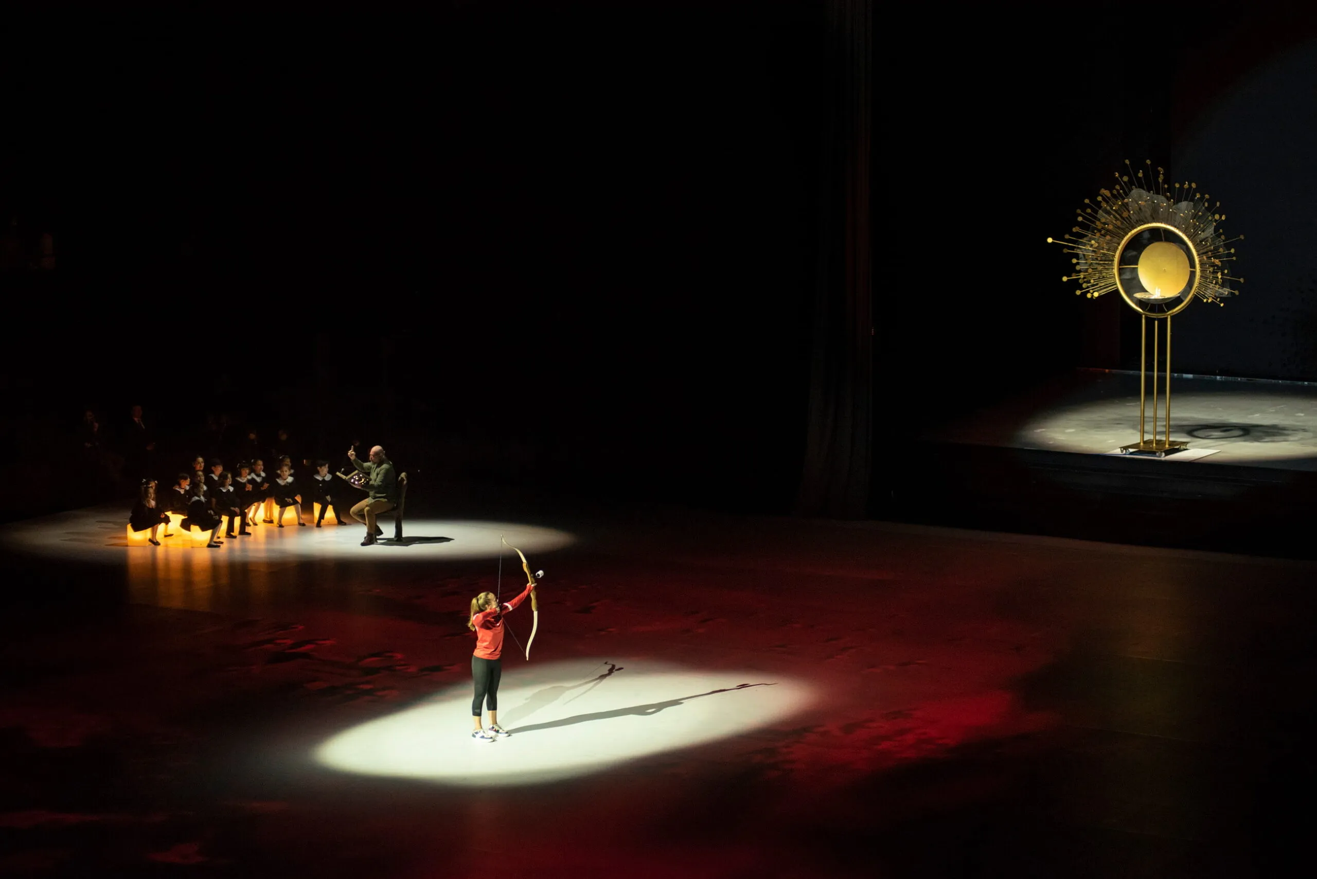 An archer in a pool of light draws her bow towards the fire sculpture across the dark arena floor