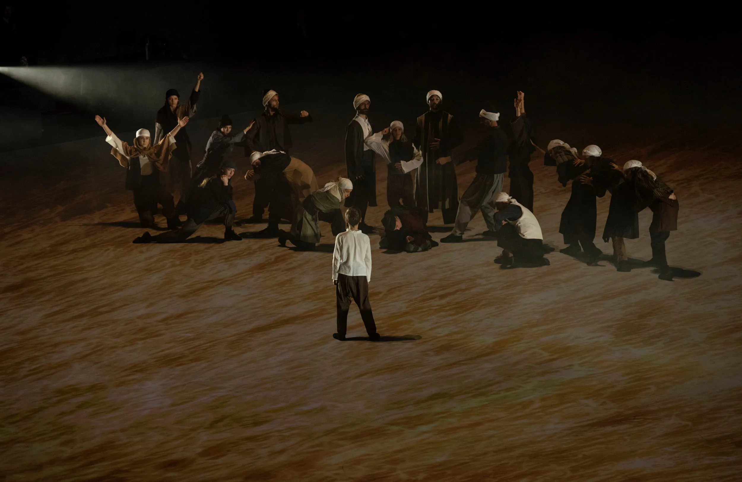 A boy in white stands alone before a group of dancers on a floor of wheat-coloured light