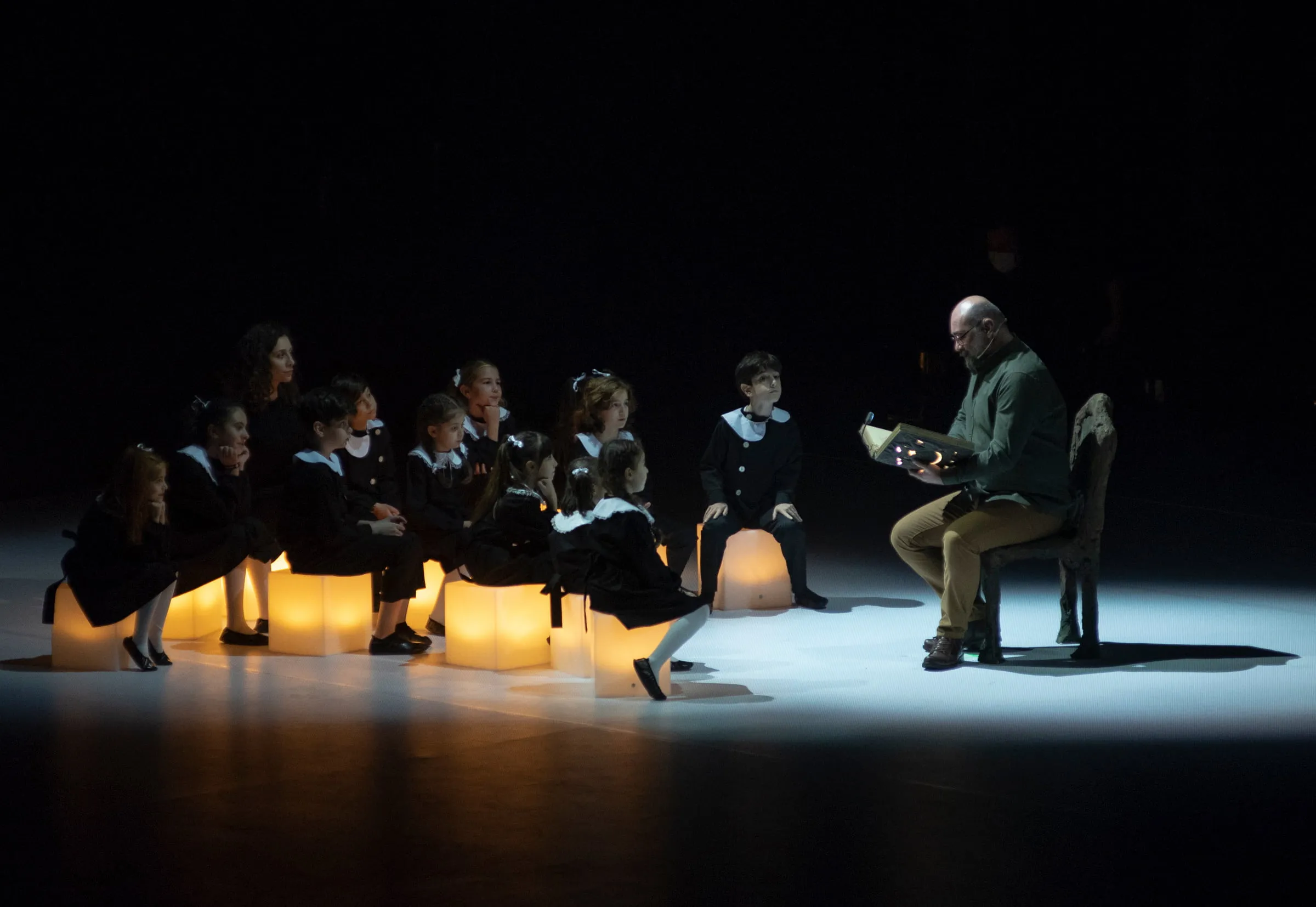 An actor in a chair reads from a book to children seated on glowing lantern cubes
