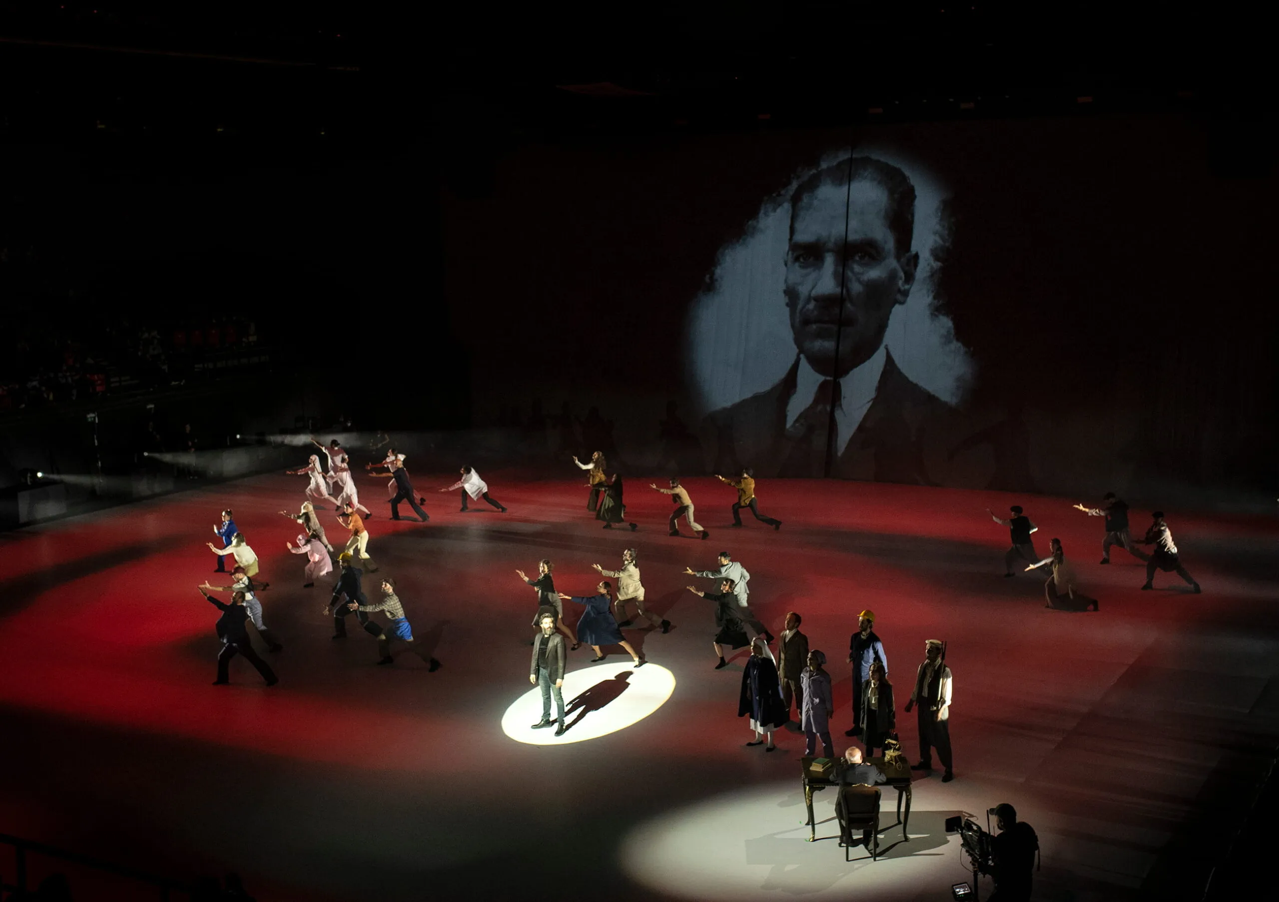 Dancers spread across a red projected floor beneath a monumental portrait of Mustafa Kemal Atatürk