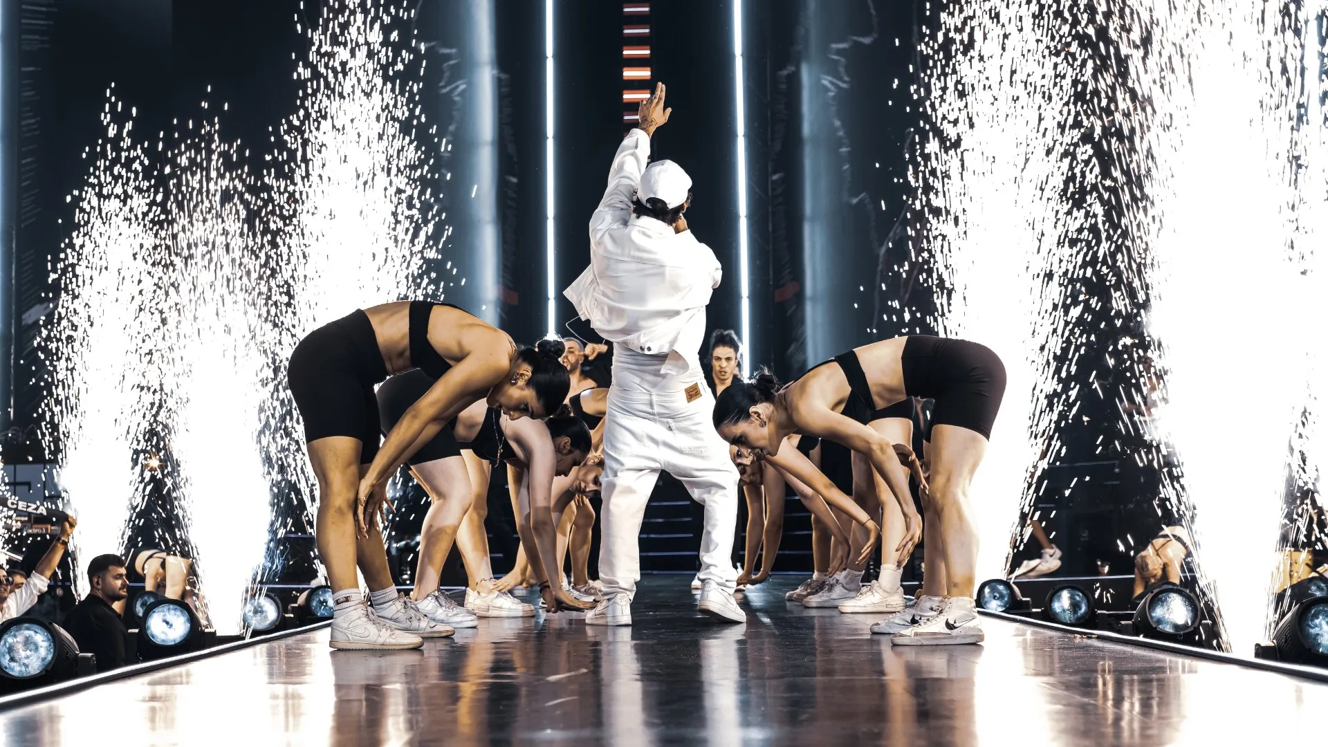 A rapper in white at the centre of crouching dancers as fountains of sparks rise behind them