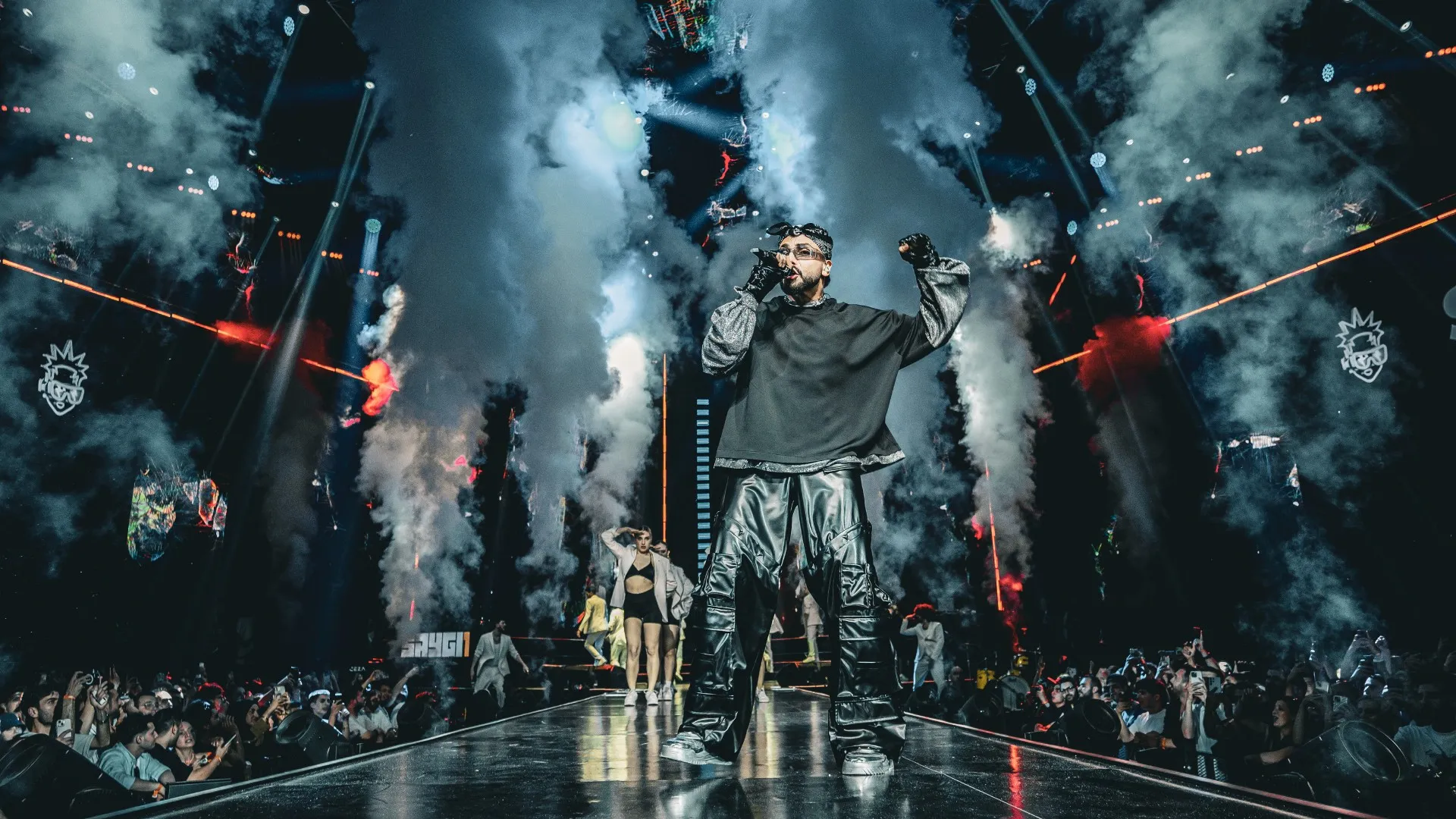 A rapper in an oversized dark jacket at the front of the stage, smoke jets firing around him