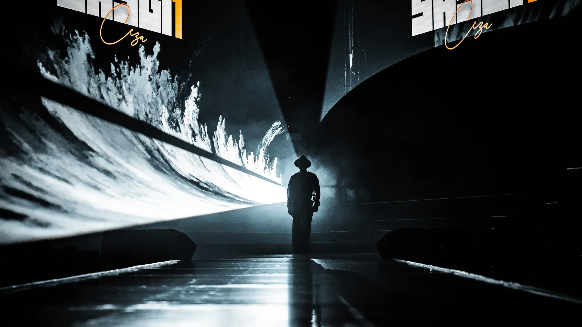 A lone figure in a hat stands at the end of the runway, white light flooding the stage behind him