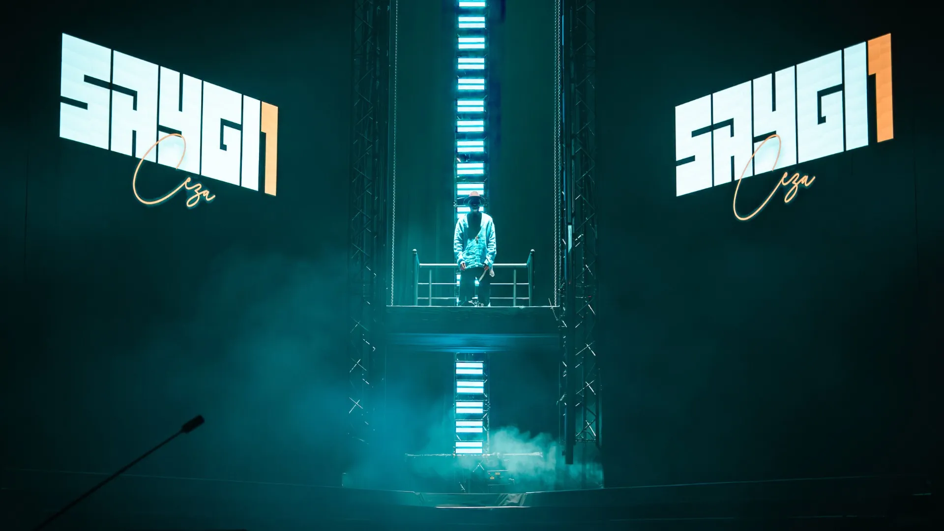 The SAYGI1 logo on two screens either side of a vertical light tower in teal haze