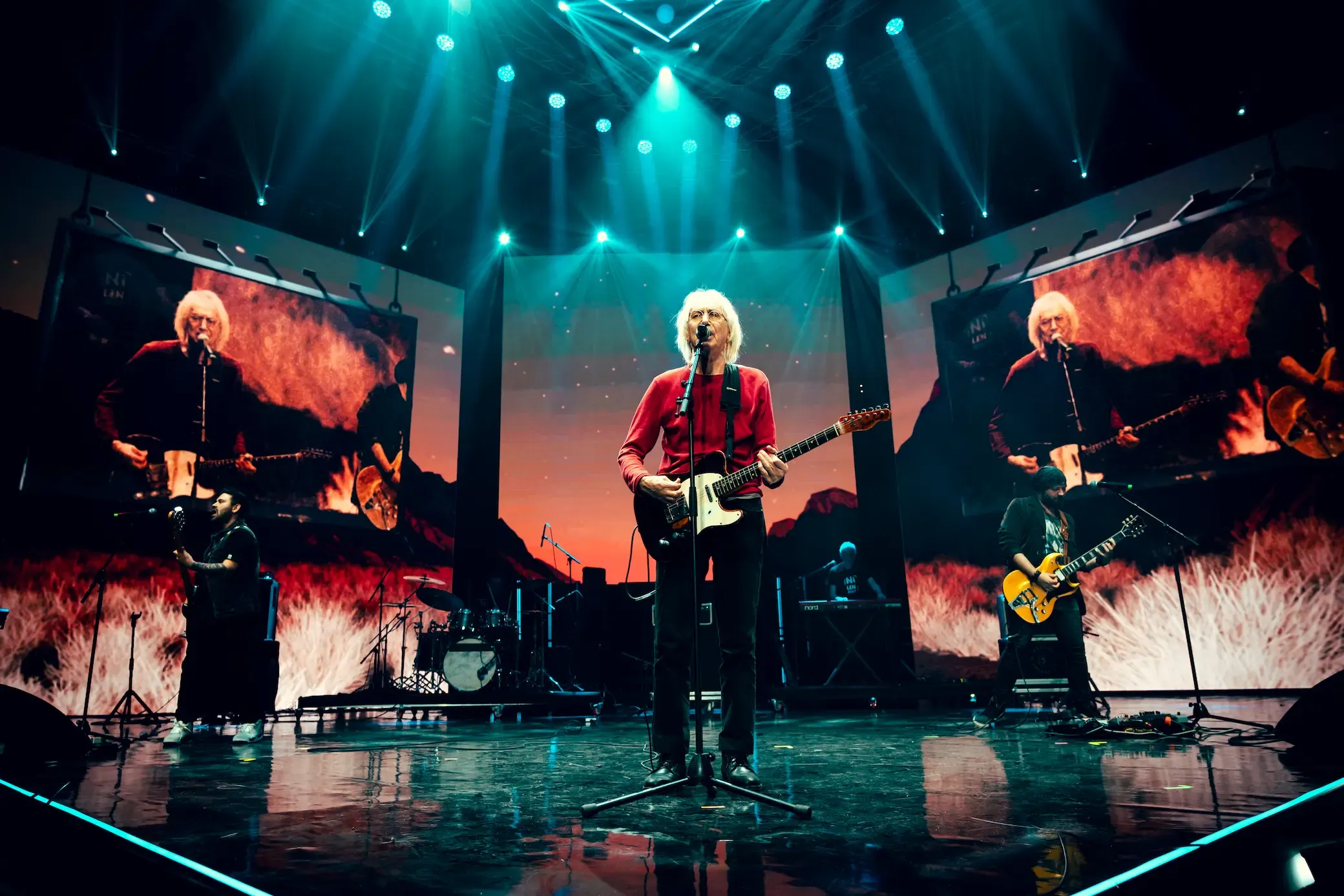 A guitarist in red at the microphone, flanked by screens of burning landscape