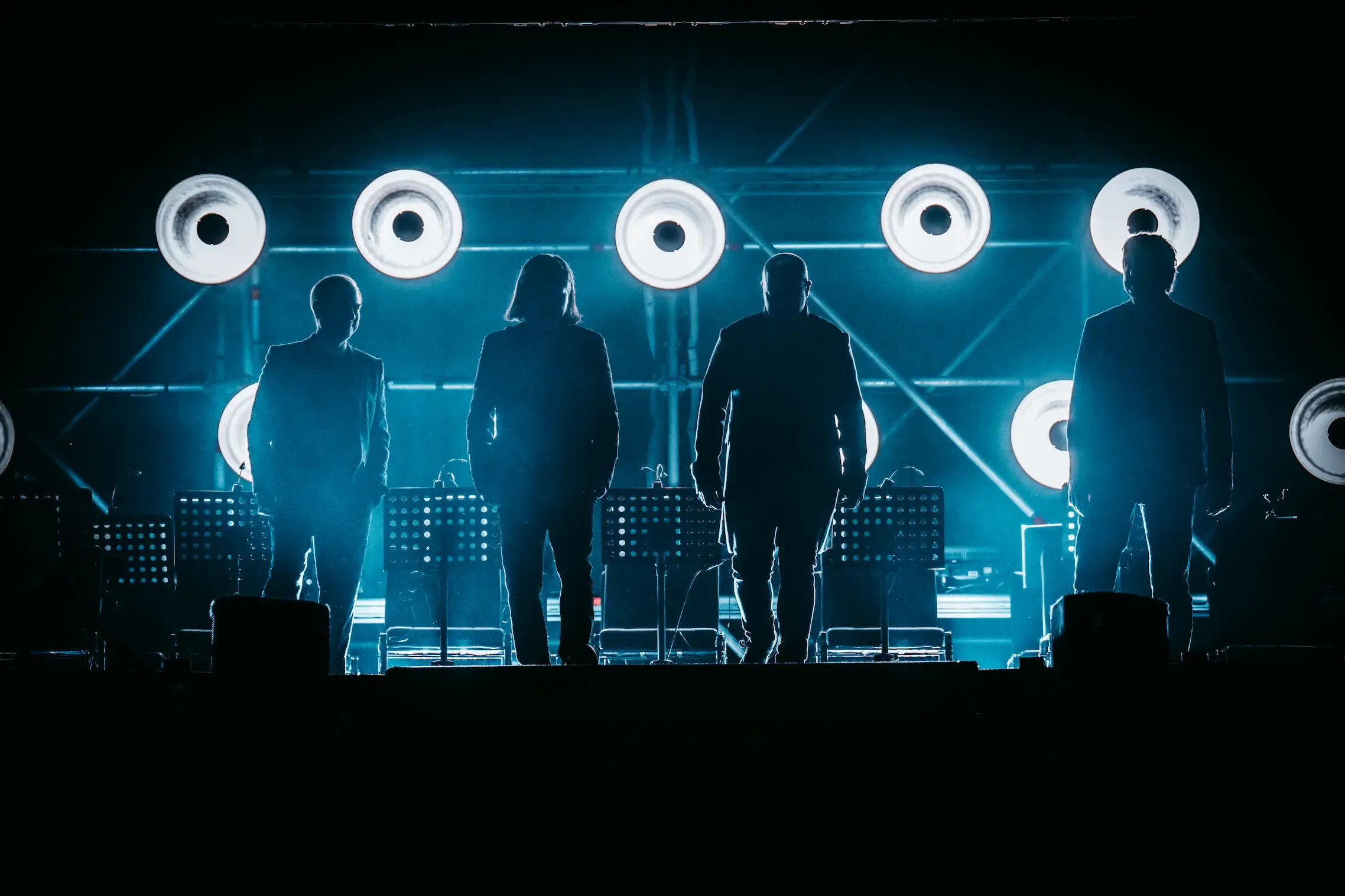 Four band members in silhouette against a row of glowing circular lights