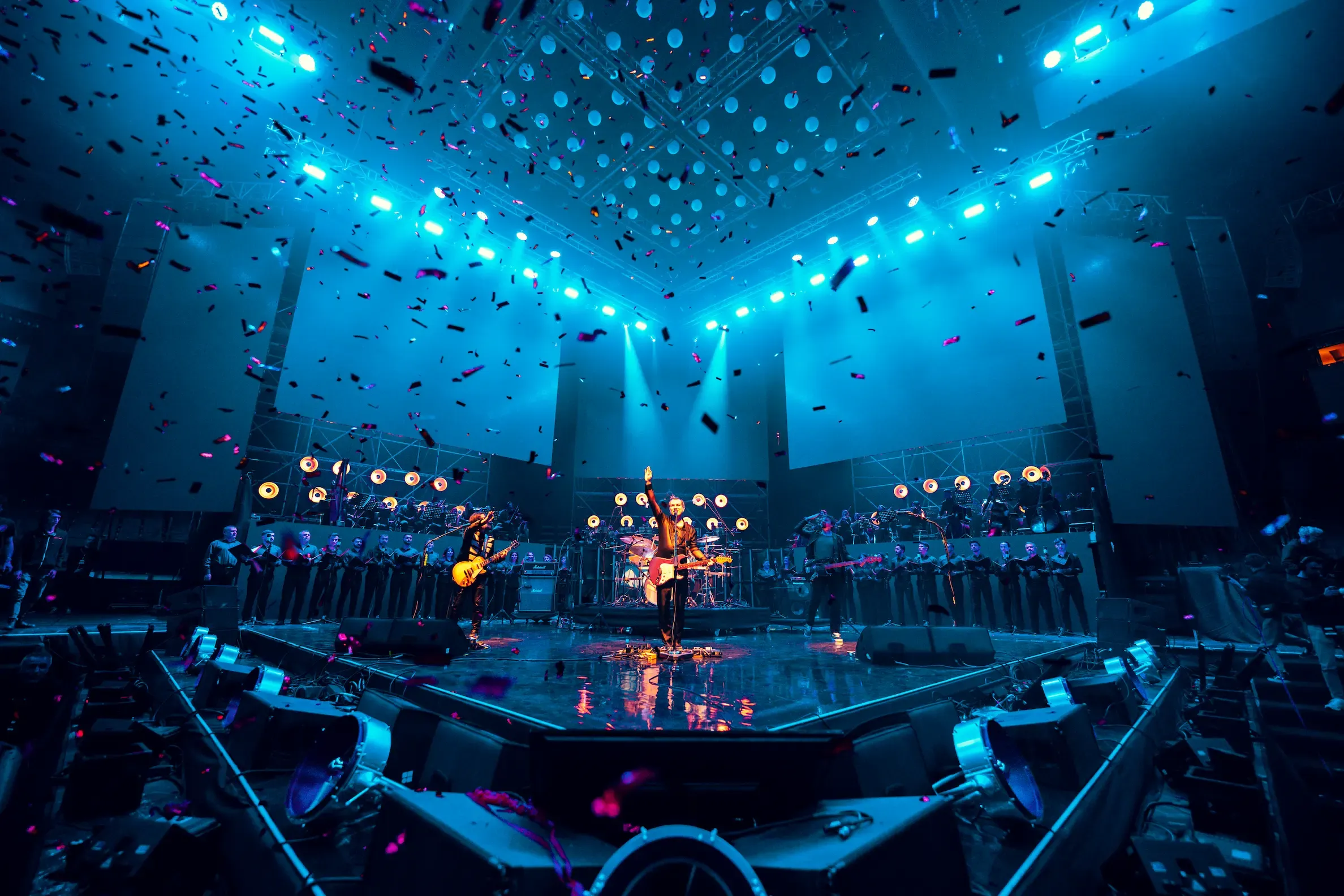 The whole arena stage with confetti falling, the orchestra on tiers either side of the band