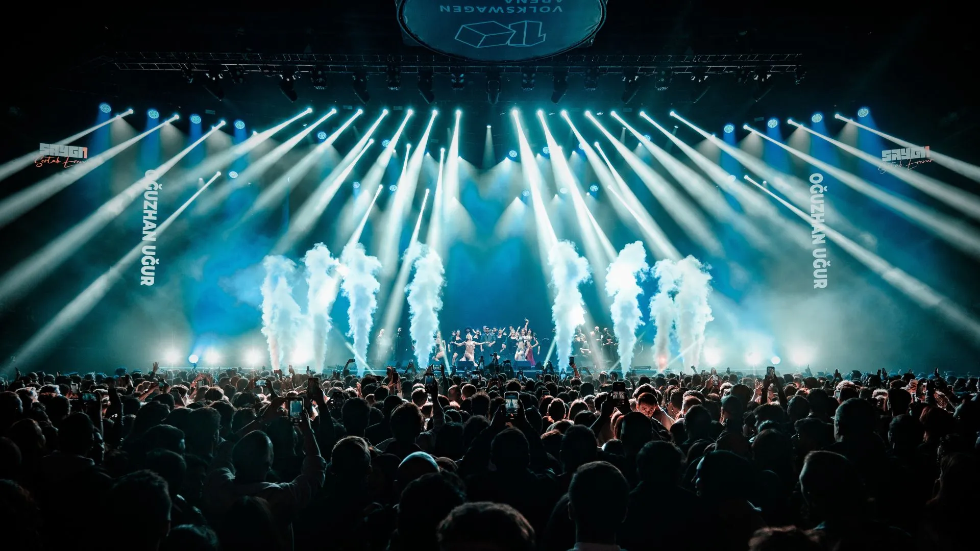 White light beams and smoke columns fan out over the stage as a full arena watches