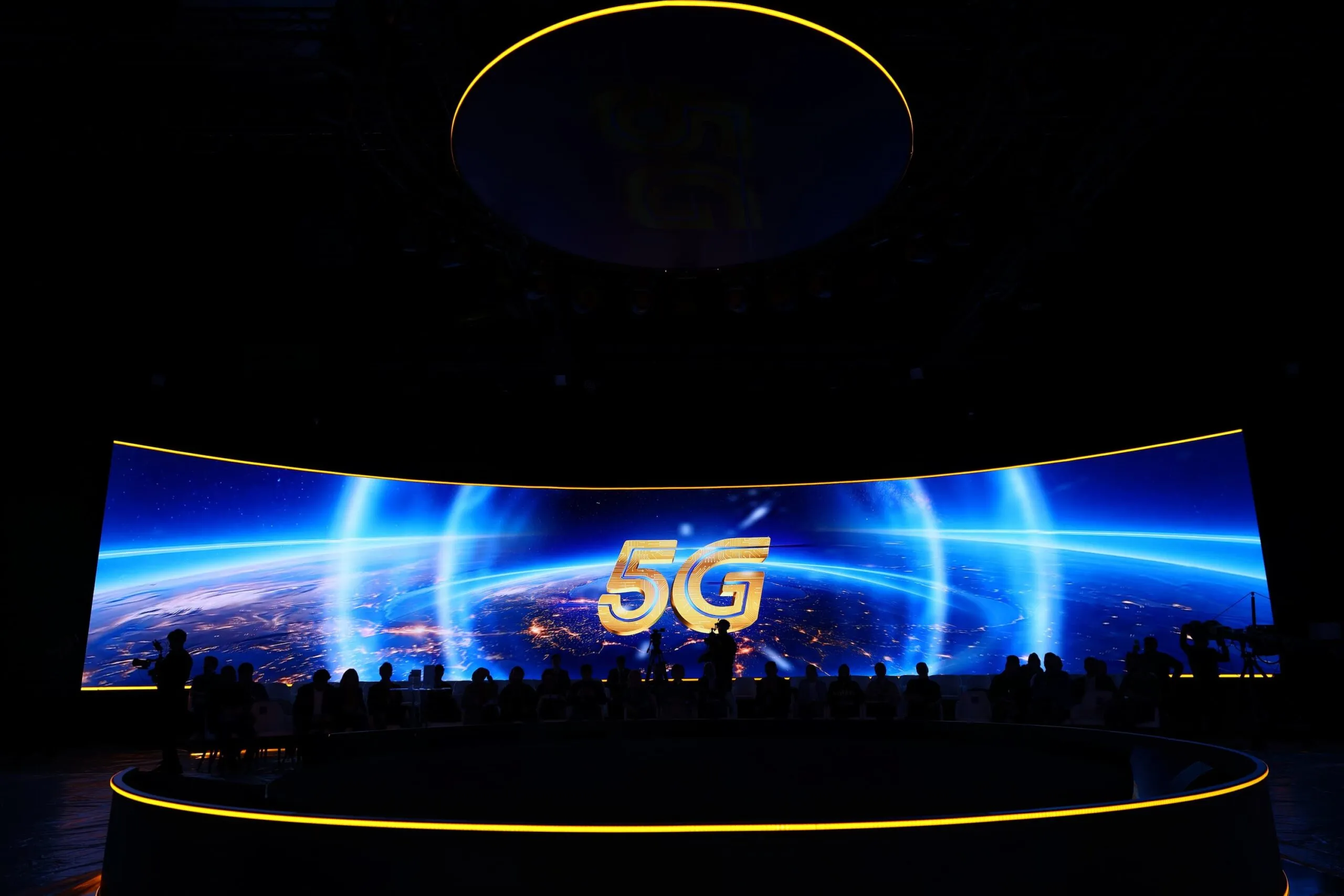 A curved LED wall showing Earth from orbit and a glowing 5G mark, the audience in silhouette below