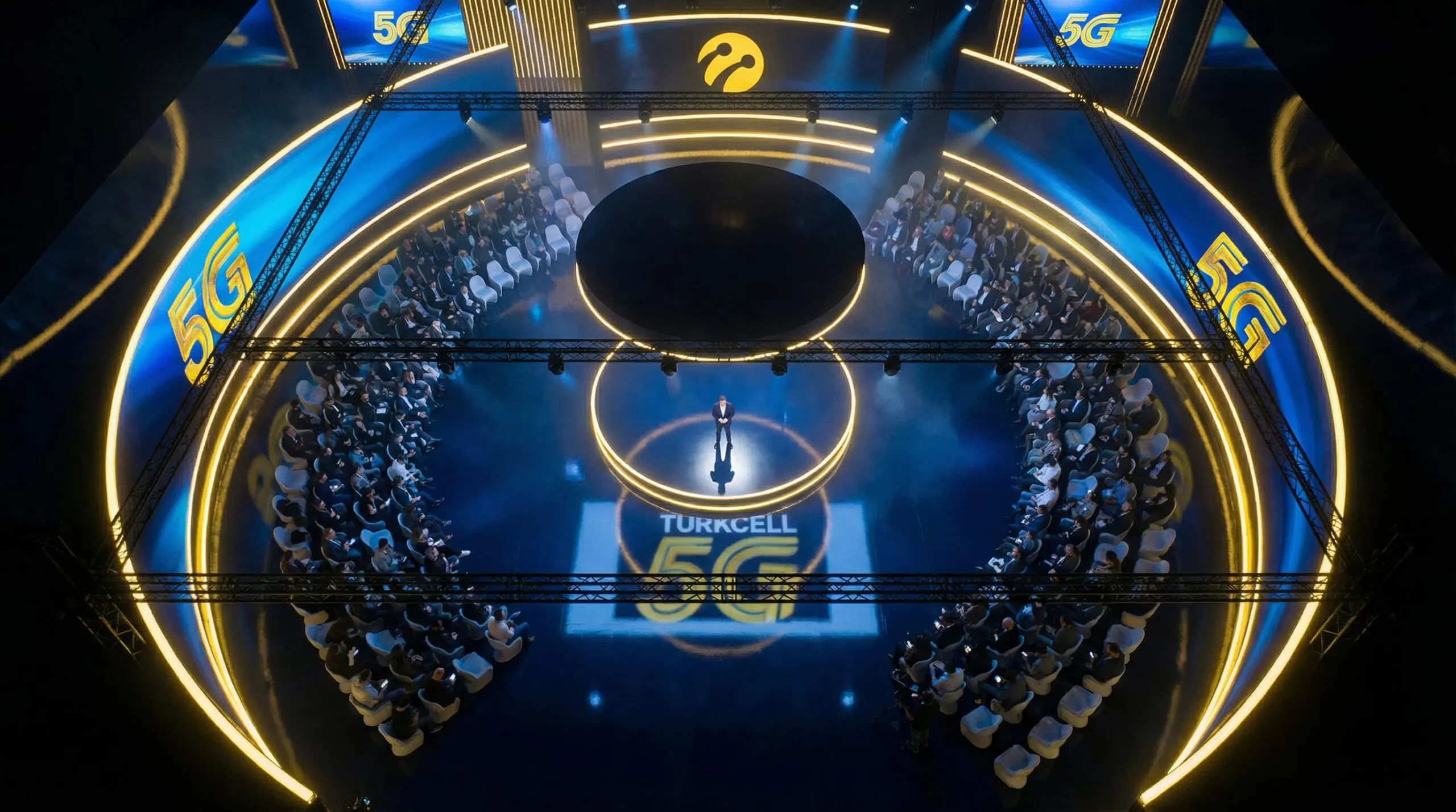 Overhead view of the 360° launch room: the audience seated in a ring around the central podium, framed by curved LED walls and gold light lines