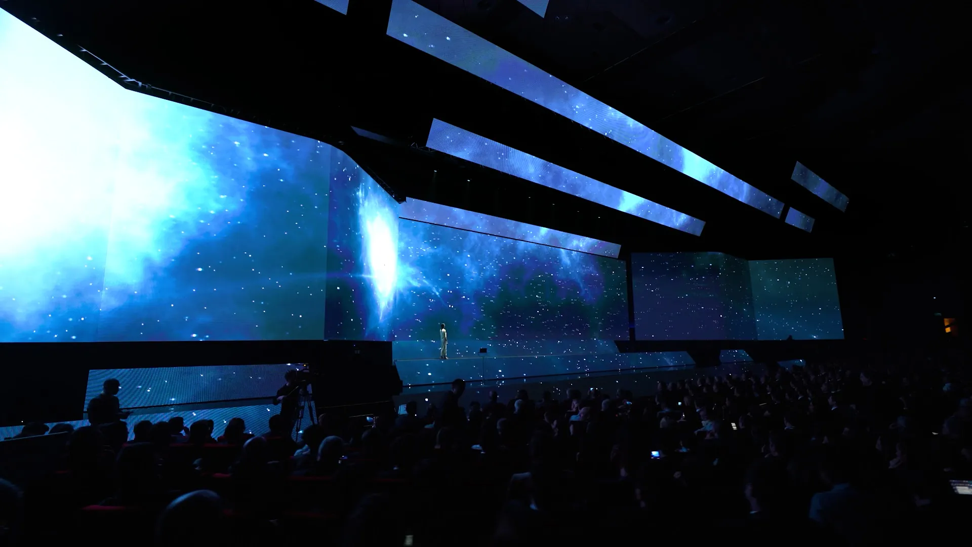 Angled LED blades sweep across the auditorium towards a vast blue screen, the audience below