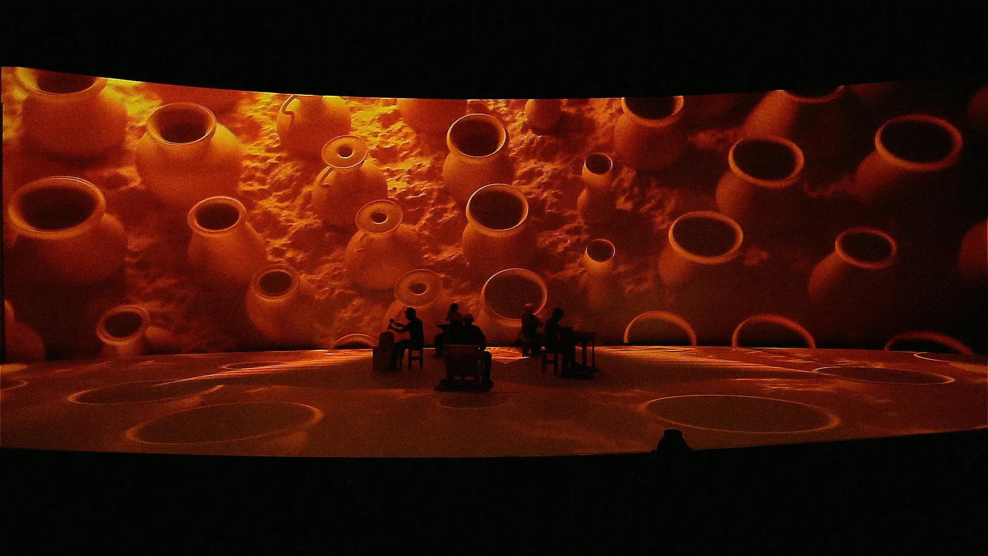 Performers seated at tables inside a projection of molten orange that wraps the whole stage