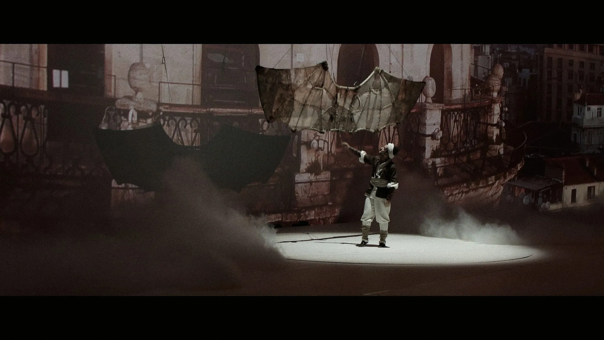 A lone figure in a spotlight raises his arm as hand-built wings hang above the stage