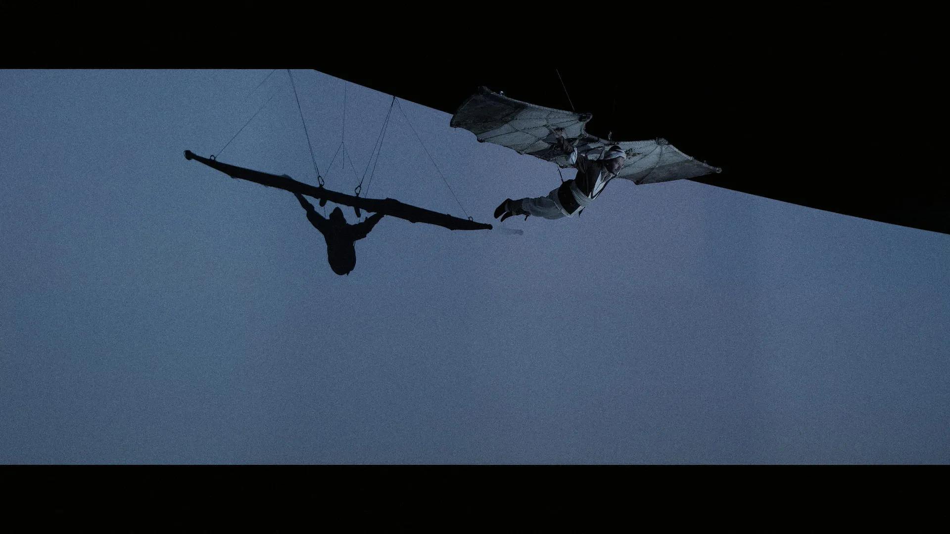 Two aerial performers flying on wings against a pale blue sky